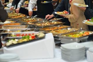 line of people on both sides of a corporate event, serving their plates along the line of food trays displayed. Desserts, vegetables and meats are being served. Pasta are being scooped by silverware. A variety of nutritious foods.